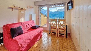 Een gezellige kamer met een rode bank, houten vloer en een tafel met stoelen. Een televisie is aan de muur bevestigd, en een raam biedt uitzicht op de bergen.