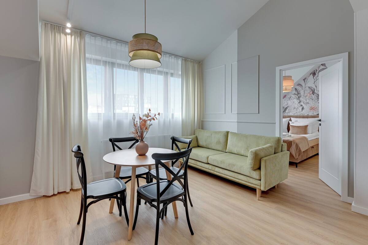 A modern living room with a dining table, chairs, a sofa, and a large window with sheer curtains.