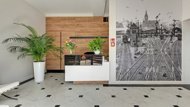 A reception area with a wooden panel wall, a reception desk, potted plants, and a large black and white photograph of a train station. - Waterlane Vintage Fitness & Parking by Downtown Apartments (Gdansk)