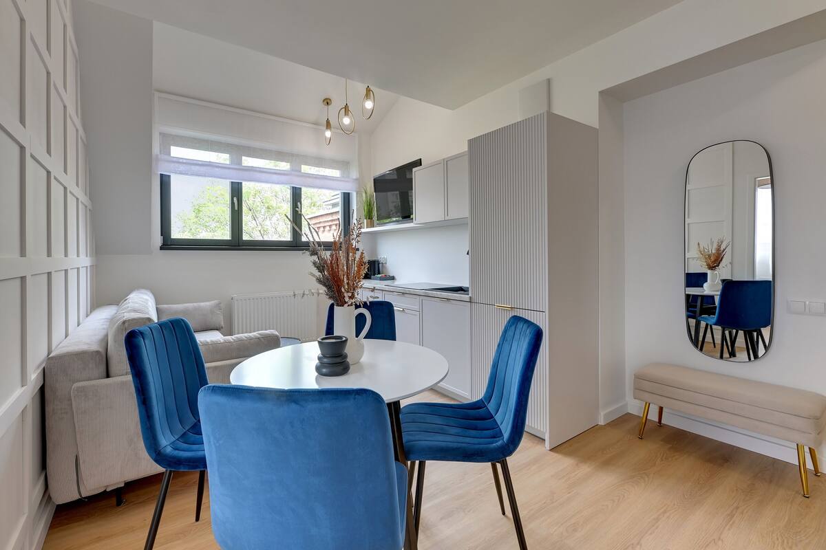 A modern kitchen with white cabinets, a dining area with a round table and blue chairs, and a large mirror on the wall.