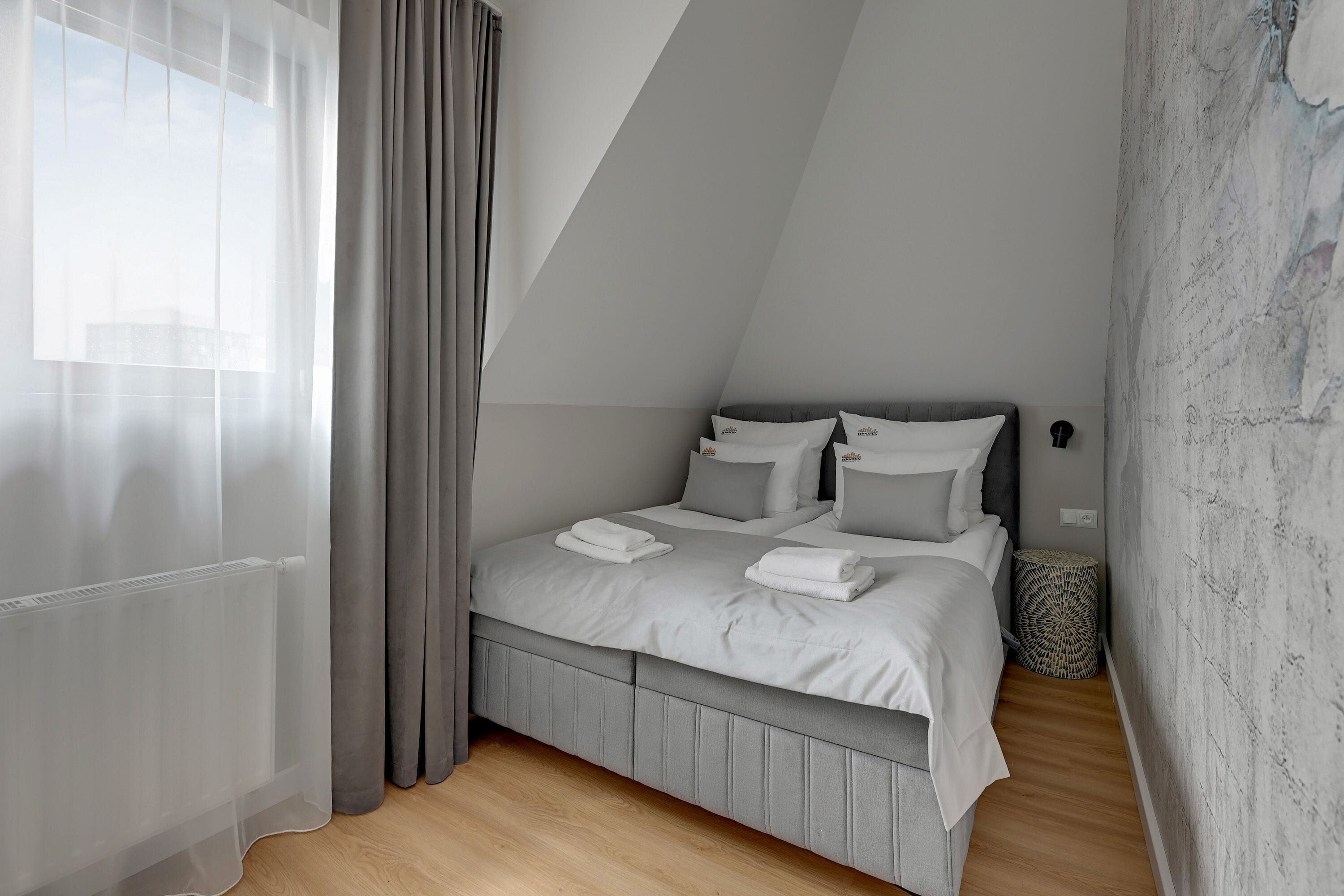 Un dormitorio moderno con techo inclinado, cama gris con ropa de cama blanca, cabecera gris y un gran ventanal con cortinas de gasa.