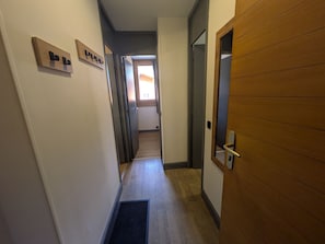 Ein schmaler Flur mit einer Holztür rechts, einer schwarzen Fußmatte am Eingang und einem kleinen Fenster am Ende.