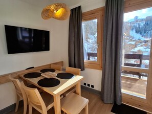 A dining area with a wooden table and chairs, a flat-screen TV mounted on the wall, and a window with curtains. - Cozy apartment in Plagne 1800, 4-5 guests, pool access (La Plagne 1800)