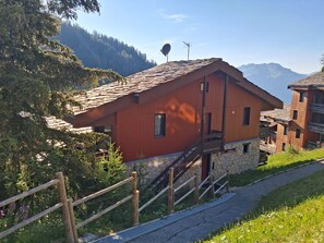 A red wooden chalet with a stone foundation, a wooden staircase, and a view of mountains in the background.