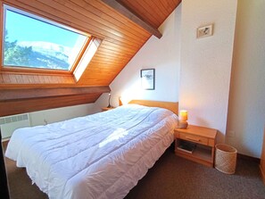 Ein Schlafzimmer mit schrägem Dach, einem Bett, einem Nachttisch, einer Lampe, einem Bild an der Wand und einem Fenster mit Bergblick.