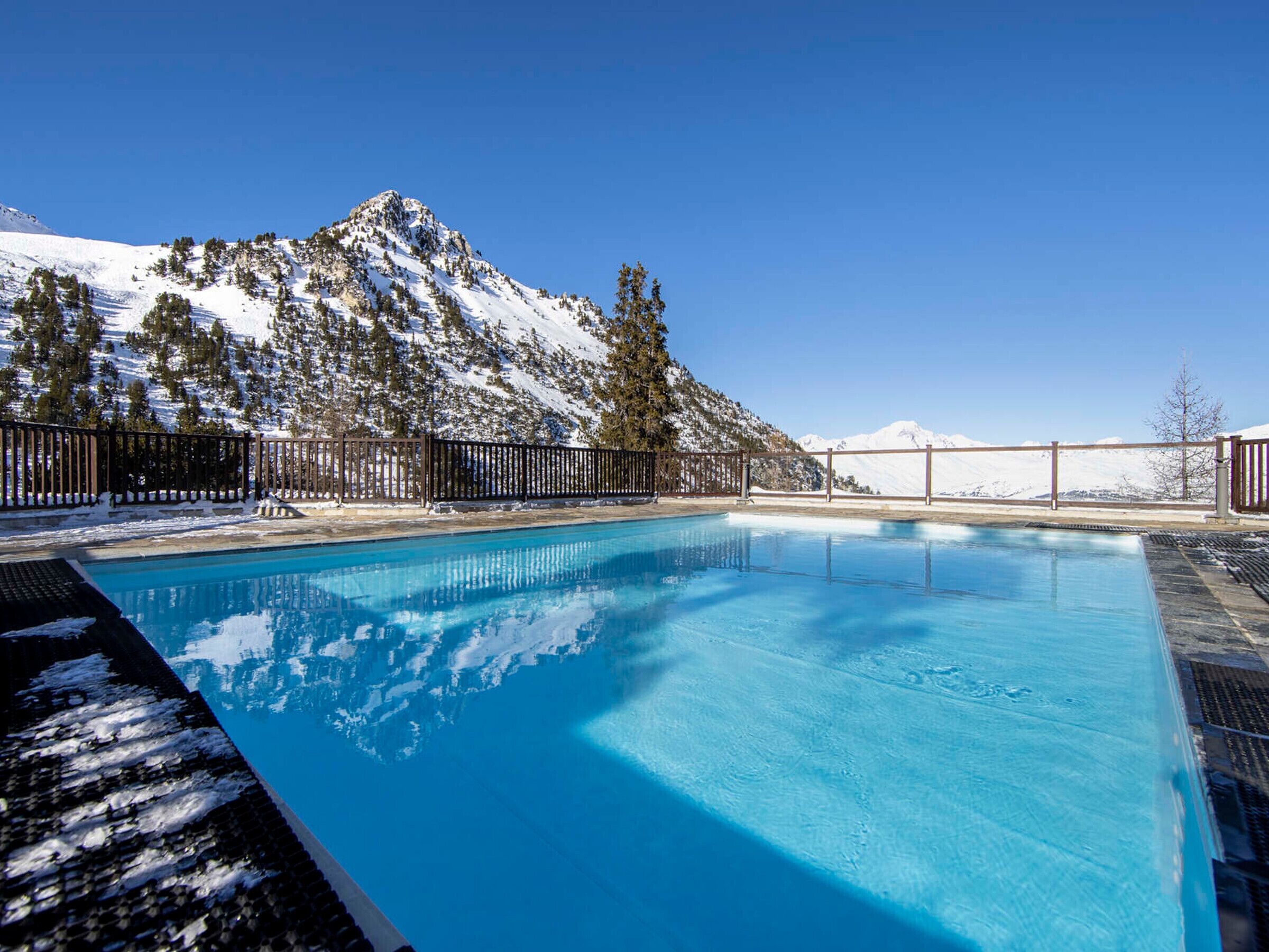 Une piscine d’un bleu éclatant, entourée d’une terrasse en bois, avec des montagnes enneigées en arrière-plan.