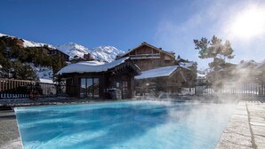 Une piscine d’un bleu éclatant, bordée d’une terrasse en bois, entourée de montagnes enneigées et d’un bâtiment de style chalet.