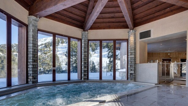 Un vaste espace intérieur abritant une piscine, agrémenté de piliers en pierre, de grandes fenêtres offrant une vue sur les montagnes et d’un aménagement intérieur moderne.