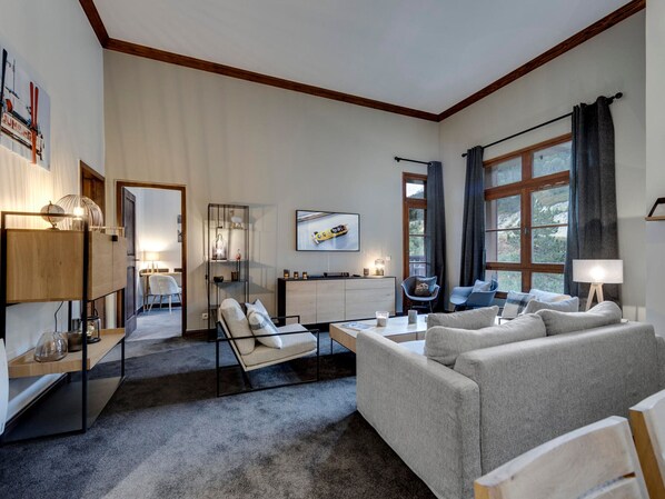 Ein modernes Wohnzimmer mit einer Couch, einem Couchtisch und einem Fernseher. Es gibt einen Essbereich mit Tisch und Stühlen. Der Raum verfügt über große Fenster mit Vorhängen.