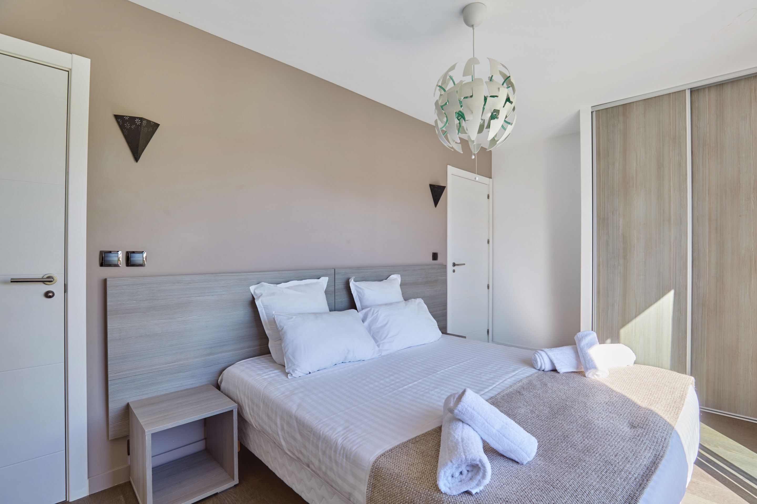 A modern hotel room with a large bed, a bedside table, a hanging light fixture, and a sliding door.