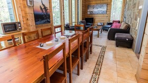 A wooden cabin interior with a dining area, a living room with a sofa and TV, and large windows.