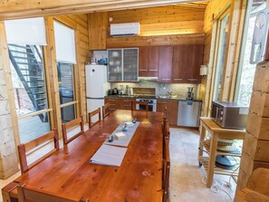 A wooden cabin interior with a dining table, chairs, a kitchen with appliances, and a window with blinds.