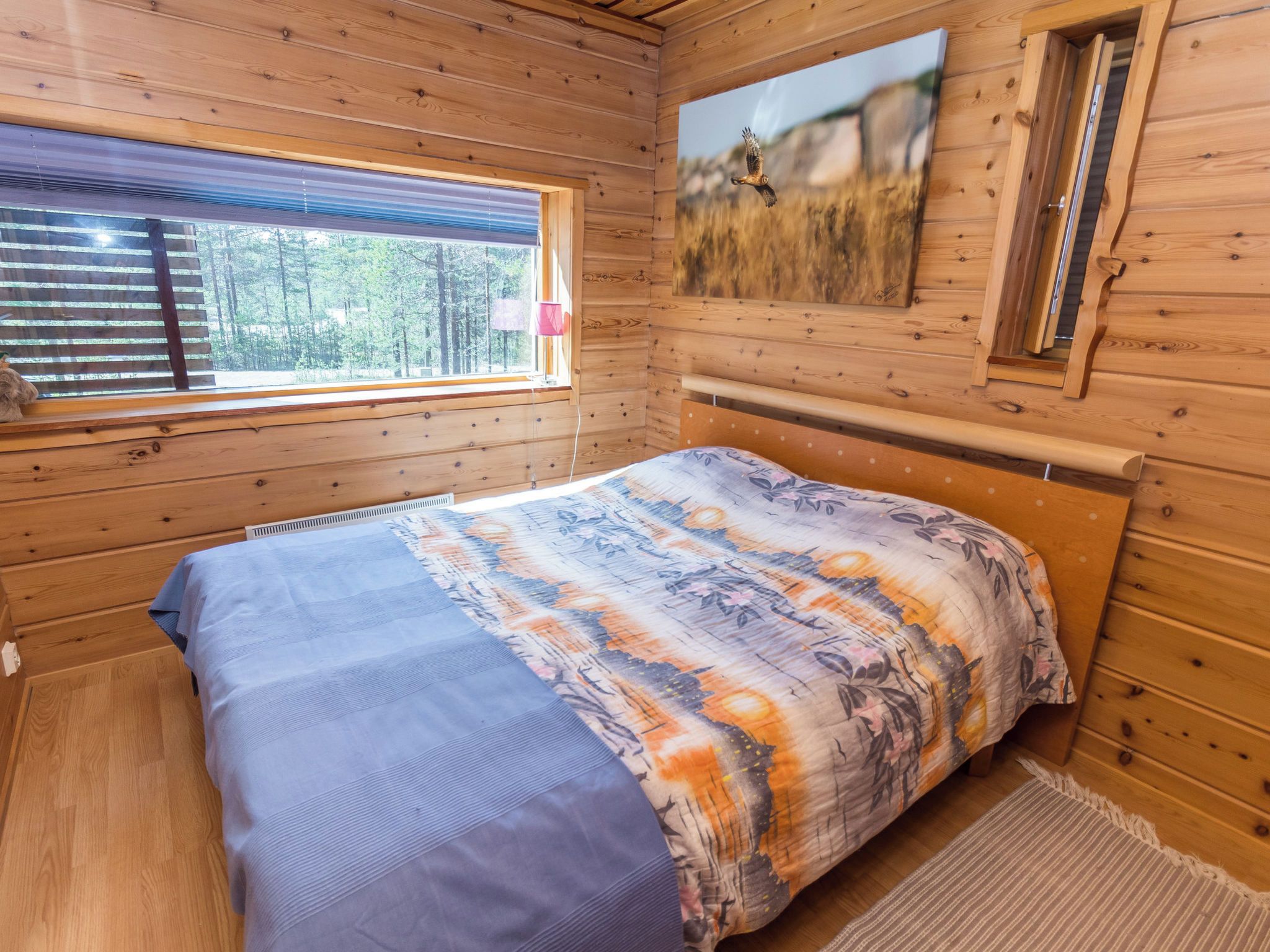 A wooden cabin bedroom with a bed, a window with blinds, a painting on the wall, and a small table with a lamp.