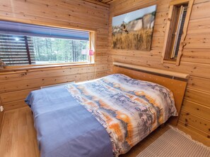 Ett sovrum i en stuga med träpanel, en säng, ett fönster med persienner, en tavla på väggen och ett litet bord med en lampa.