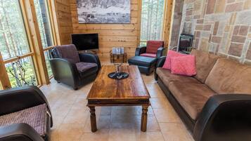 A cozy interior with a wooden floor, a stone fireplace, and a large window overlooking a forest.