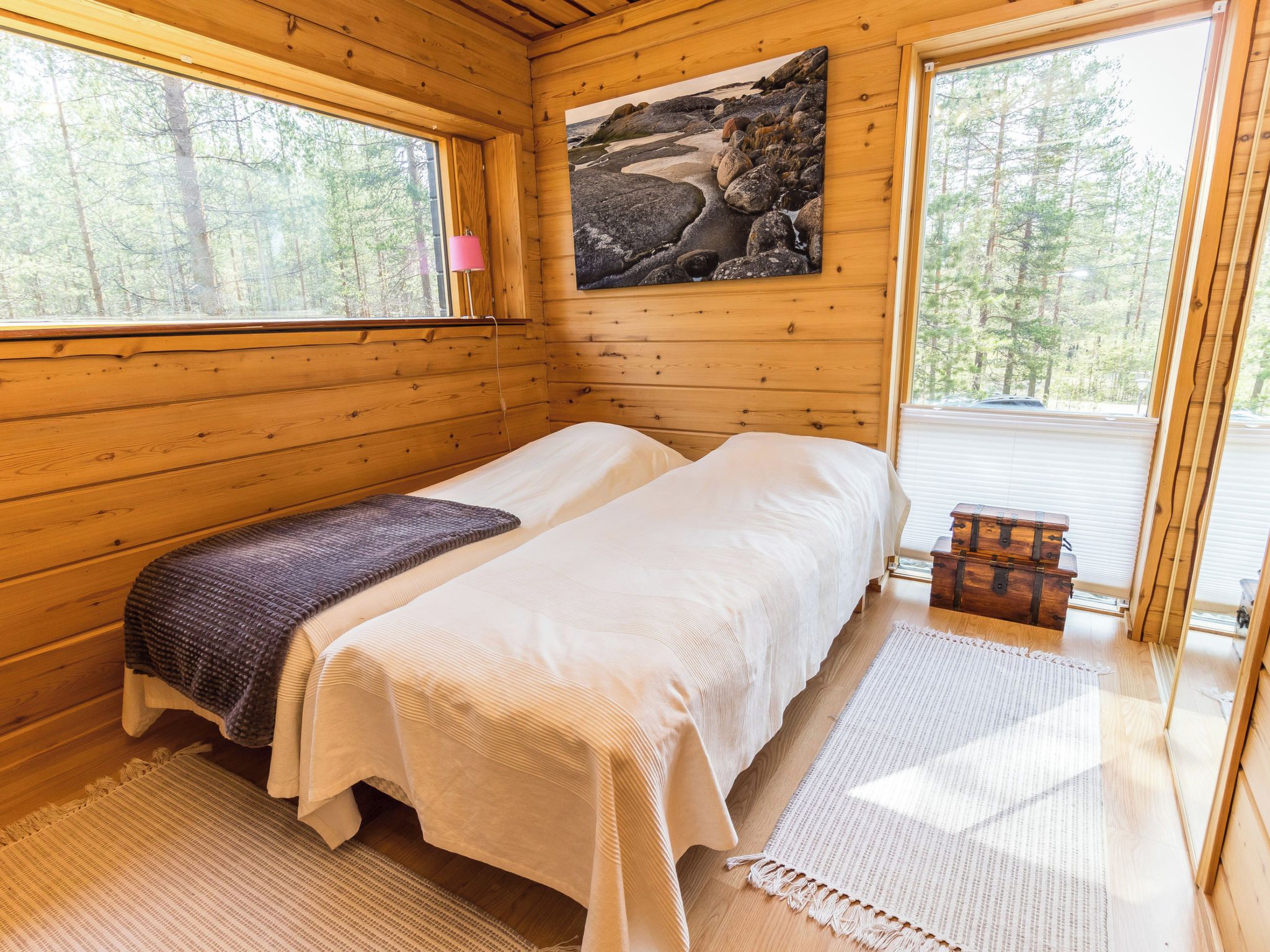 A wooden cabin bedroom with two beds, a painting of rocks, a window, and a view of trees.