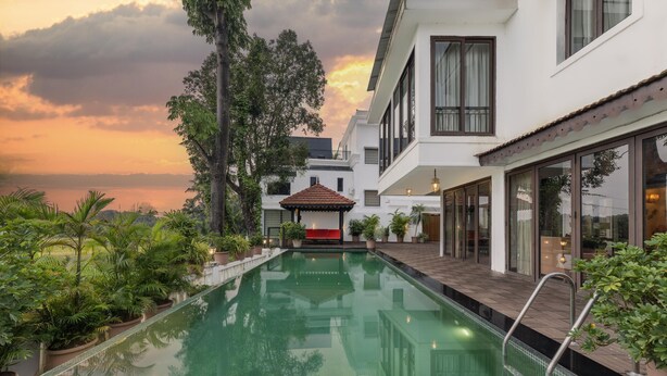 A swimming pool with a wooden deck and a gazebo, surrounded by lush greenery and modern buildings. - Echor Reis Magos Villa Goa (Verem)