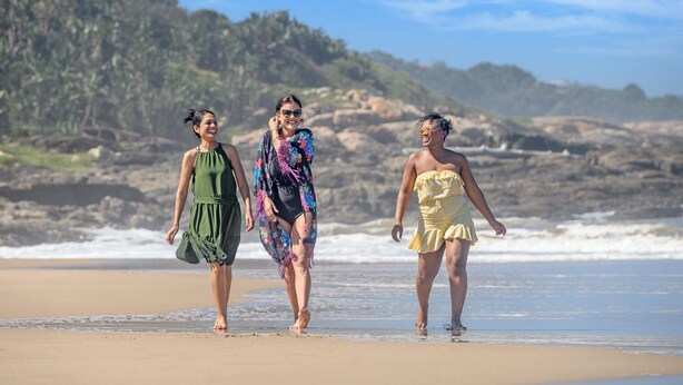 Drie mensen wandelen op een strand met rotsachtige kliffen op de achtergrond.
