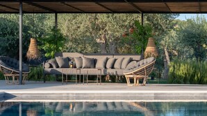 A poolside area with a sofa and chairs under a pergola, surrounded by greenery.