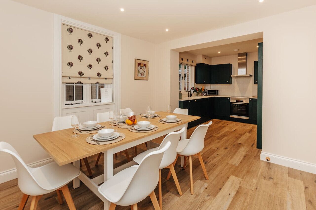 A dining area with a wooden table set for four, white chairs, and a window with a patterned blind.