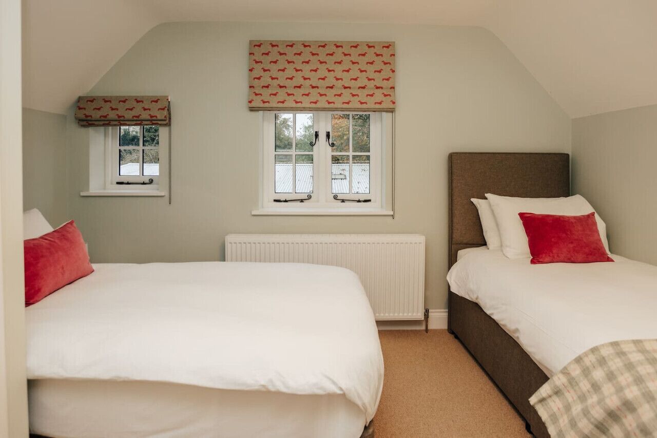A bedroom with two beds, a window with a patterned valance, a radiator, and a small window with a curtain.