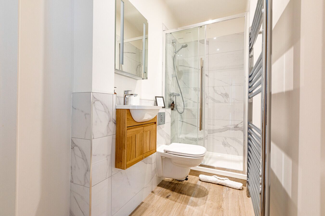 A modern bathroom with a marble-tiled wall, a wooden vanity, a white toilet, and a glass-enclosed shower area.