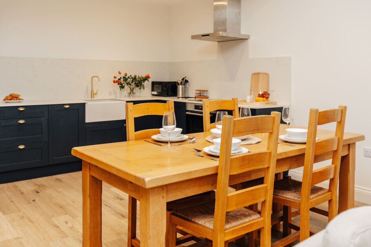 Un coin repas avec une table et des chaises en bois, un évier blanc et un four à micro-ondes à l’arrière-plan.