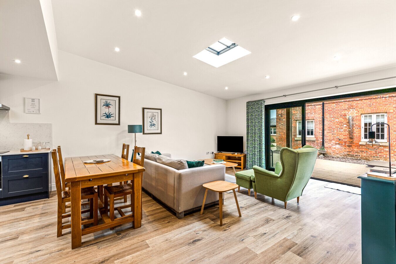 A modern living room with a wooden dining table, a sofa, and a green armchair. There is a TV and a small table with a lamp. The room has a skylight and a view of a brick building outside.