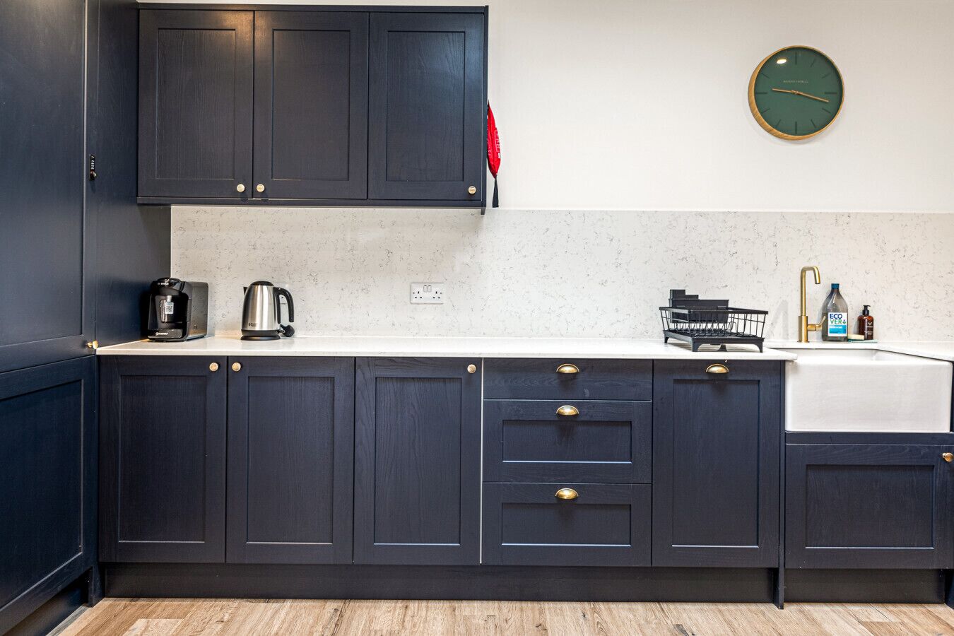 Una cocina moderna con armarios oscuros, una encimera blanca, cafetera, hervidor, pileta con grifo y un reloj de pared.