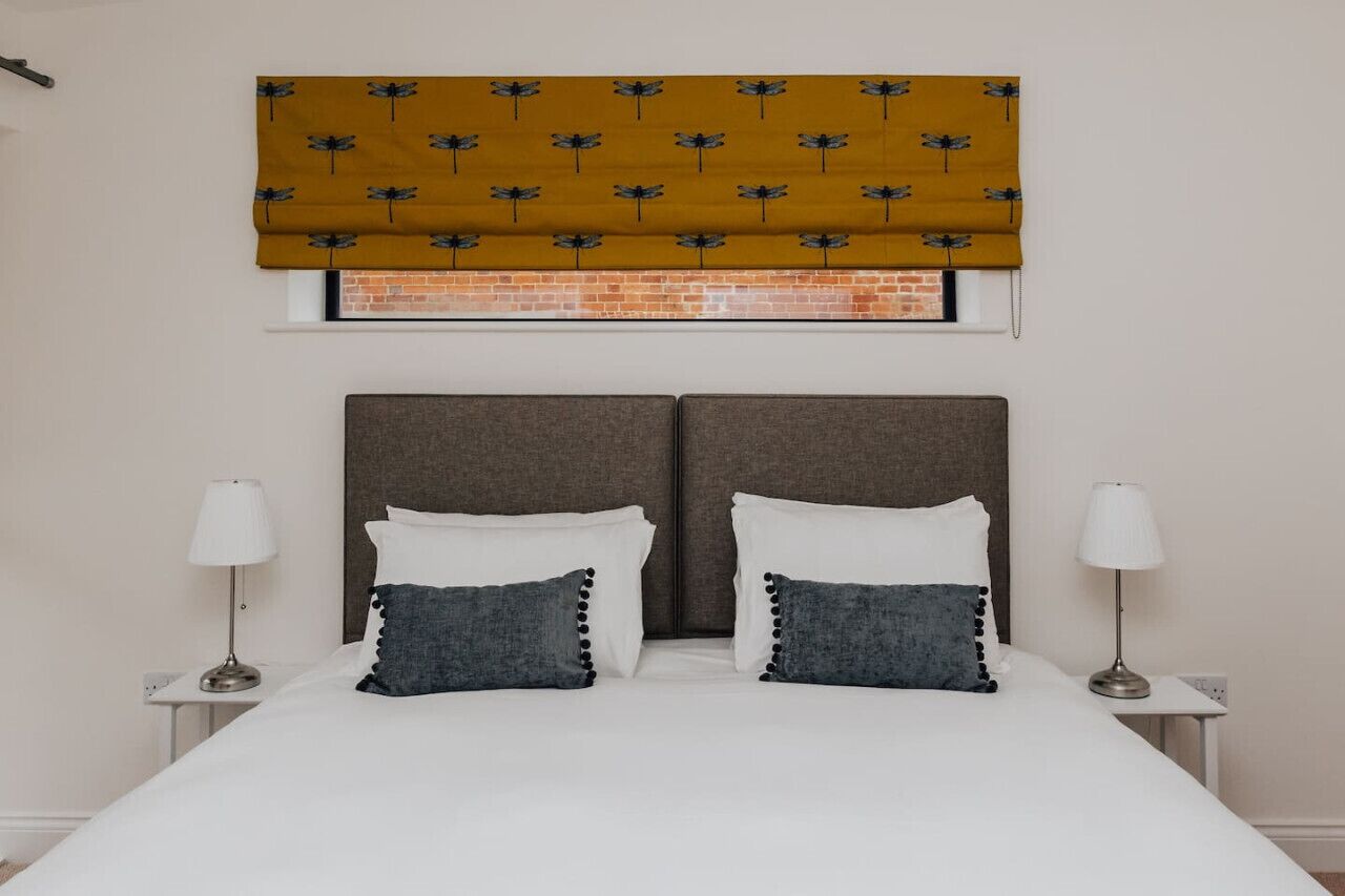 Quarto com uma cama de casal, duas mesas de cabeceira com luminárias, cabeceira e janela com persiana de rolo marrom.
