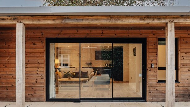 Ein modernes Gebäude mit Holzfassade und großen Glasfenstern, die in einen Innenraum mit einer Sitzecke und einem Essbereich führen.