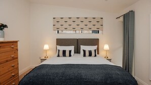 Ein Schlafzimmer mit einem Bett, zwei Nachttischlampen, einer Holzkommode, einem gemusterten Kopfteil und einem Fenster mit Vorhängen.