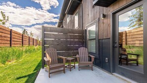 Un patio con due poltrone Adirondack e un tavolino, una recinzione in legno e una casa con un'ampia porta finestra.