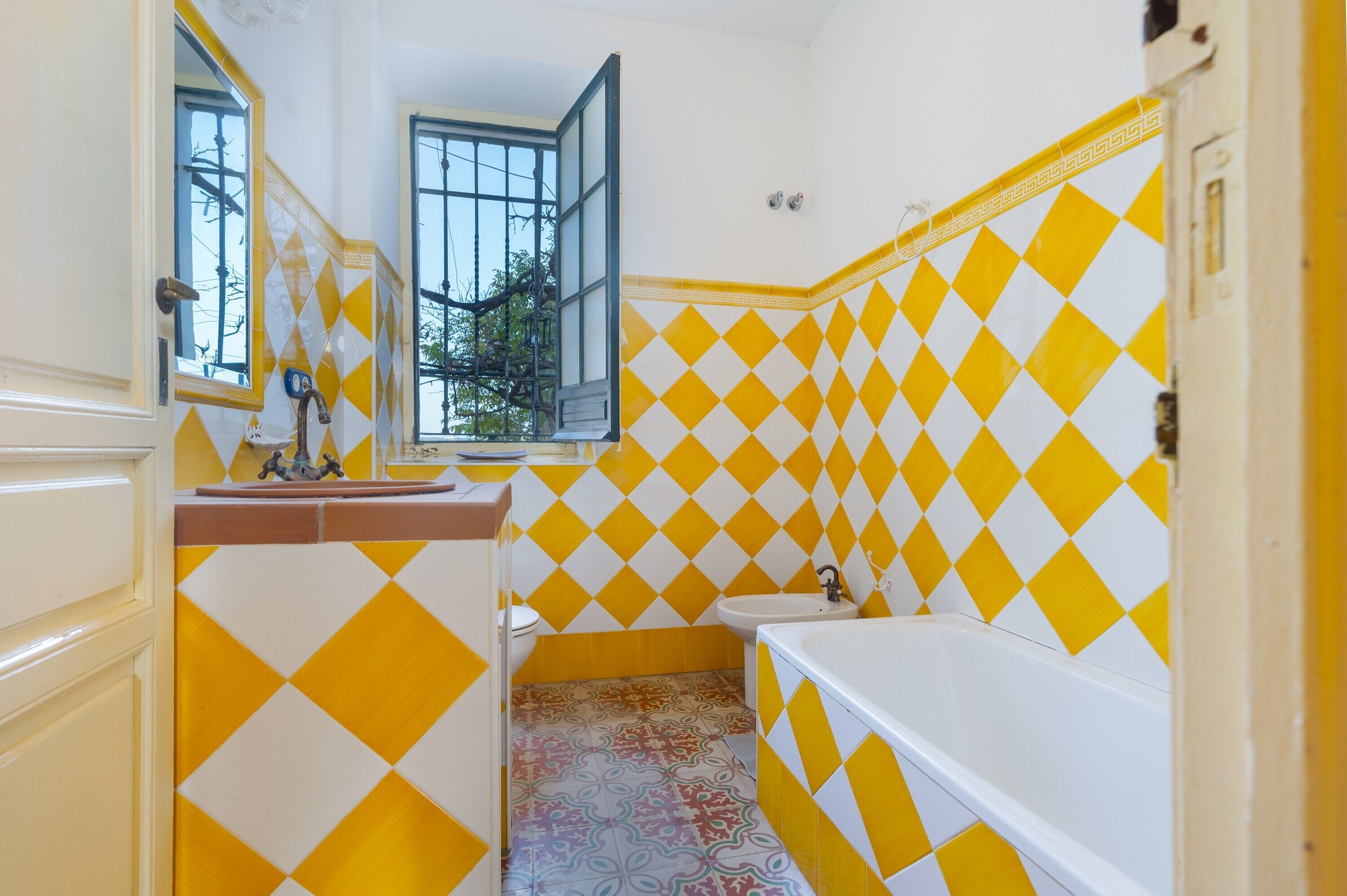 A bathroom with yellow and white diamond-patterned tiles, a white bathtub, a sink, and a window with an open shutter.