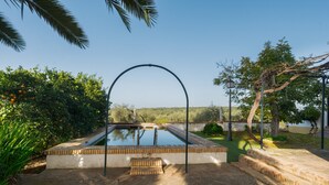 A rectangular pool with a metal arch, surrounded by greenery and a clear sky.