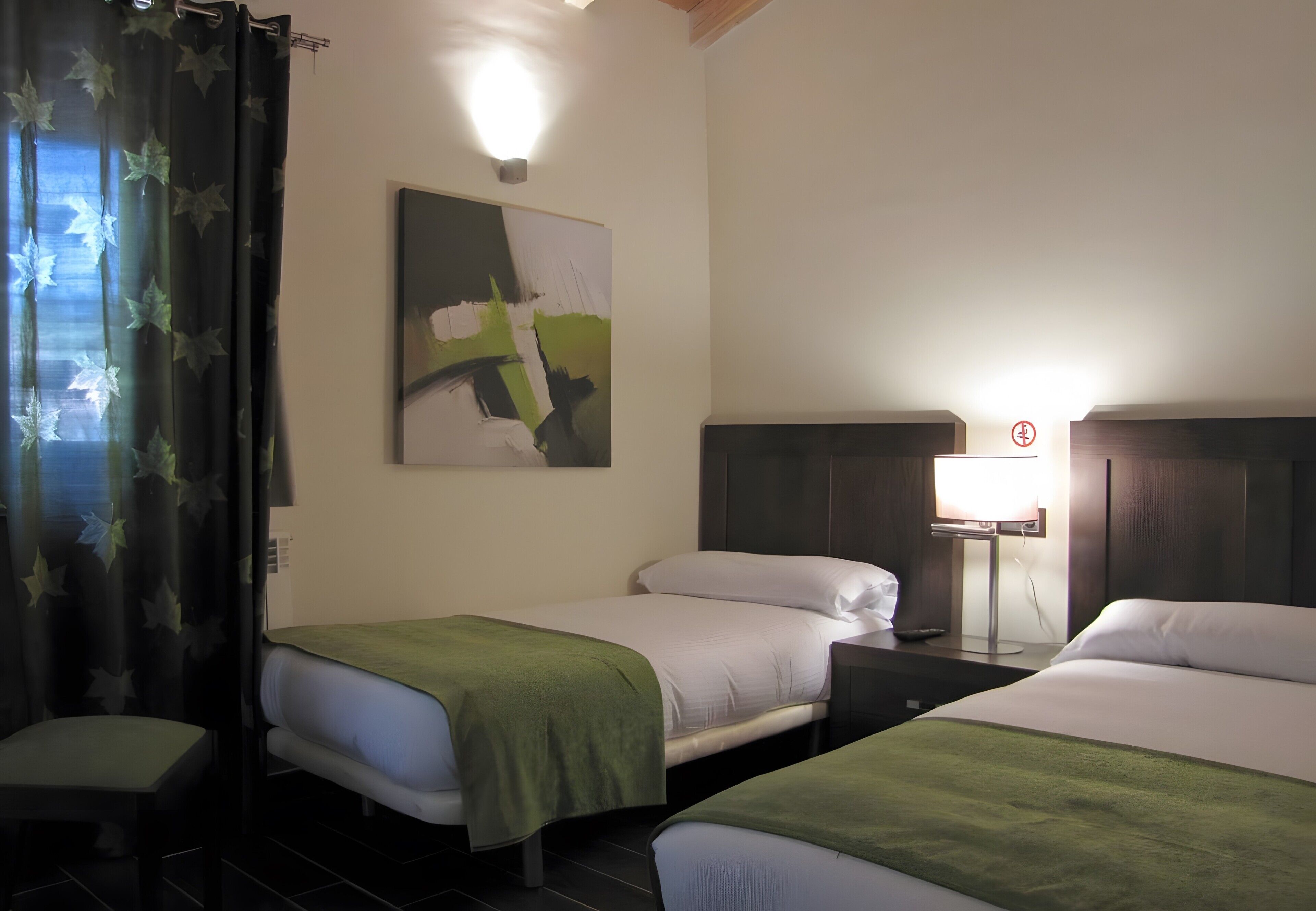 Quarto de hotel com duas camas, uma mesa de cabeceira, um abajur, um quadro na parede e uma janela com cortinas.