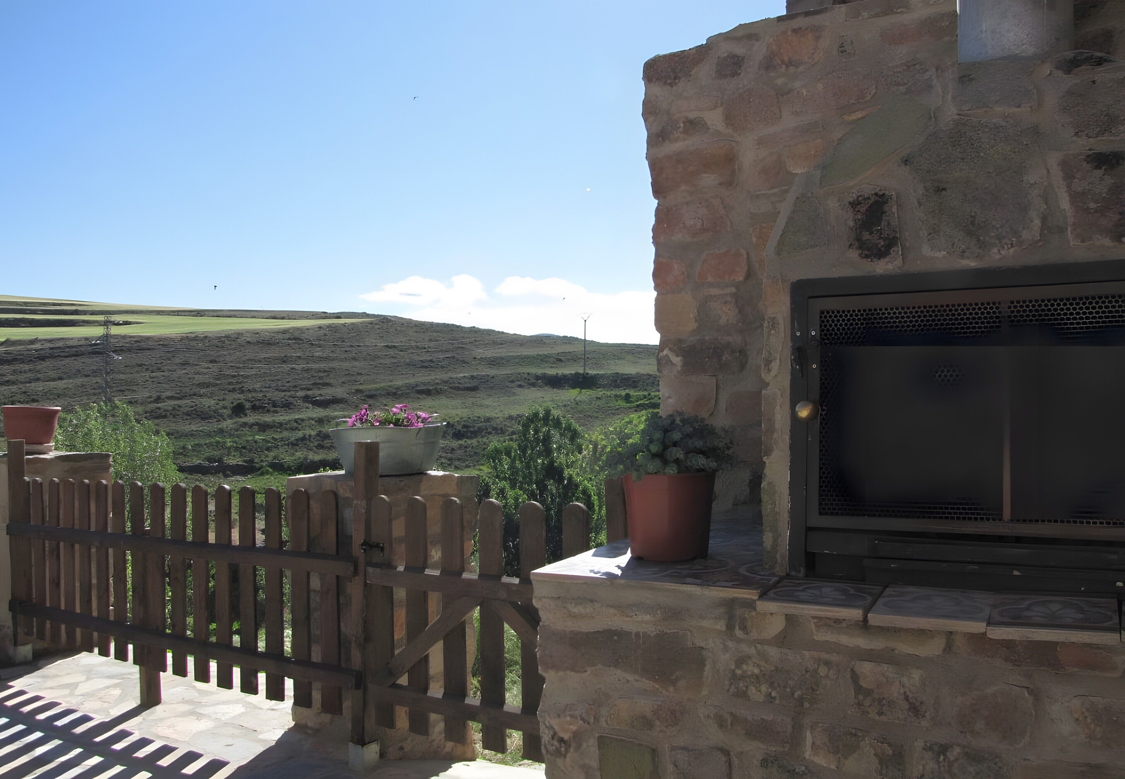 Un edificio de piedra con una parrilla, plantas en macetas y una valla de madera con vistas a un paisaje montañoso.