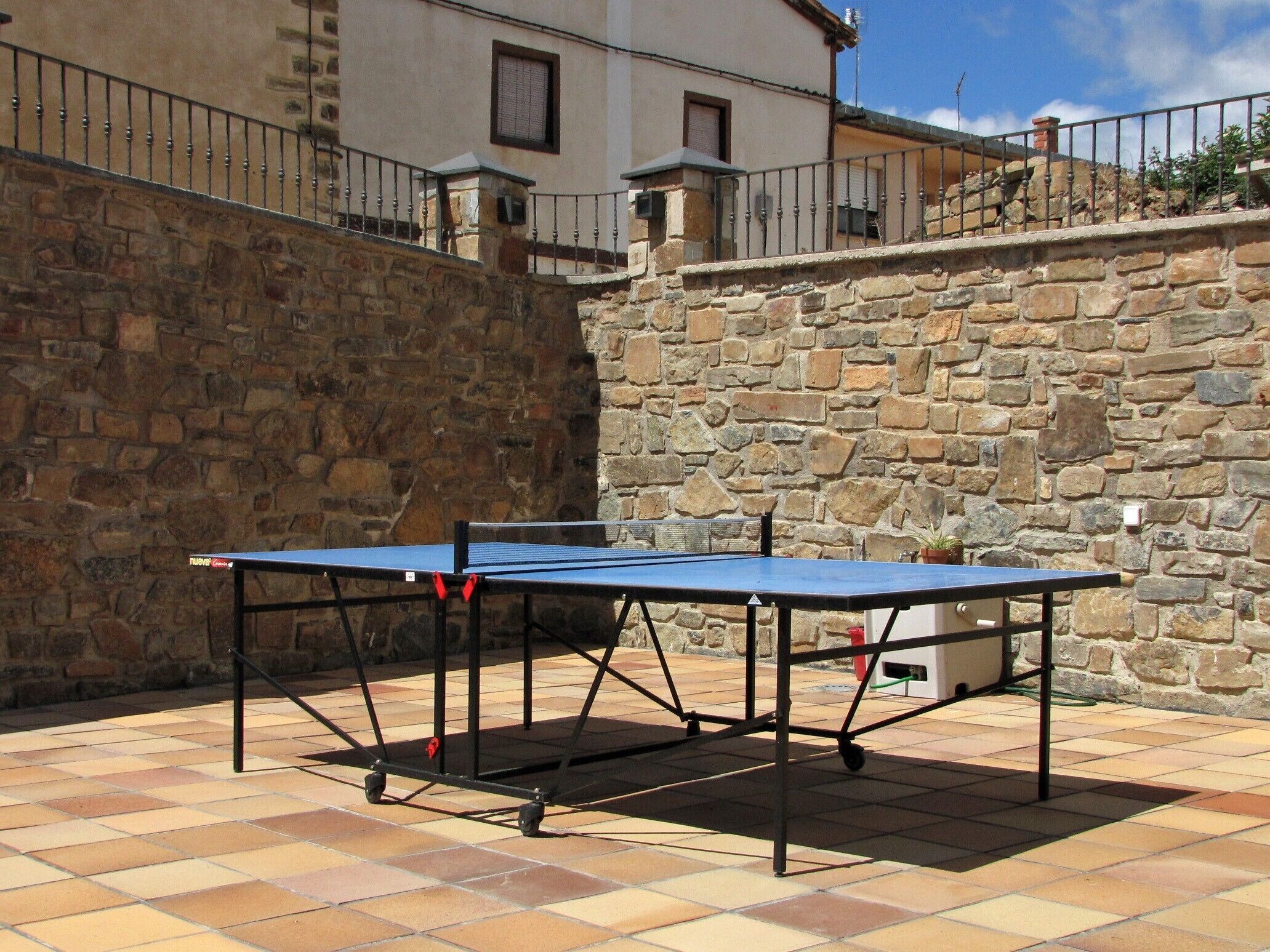 Et bordtennisbord er placeret udendørs på en fliseterrasse med en stenvæg og bygninger i baggrunden.