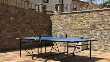 Et bordtennisbord er placeret udendørs på en fliseterrasse med en stenvæg og bygninger i baggrunden.