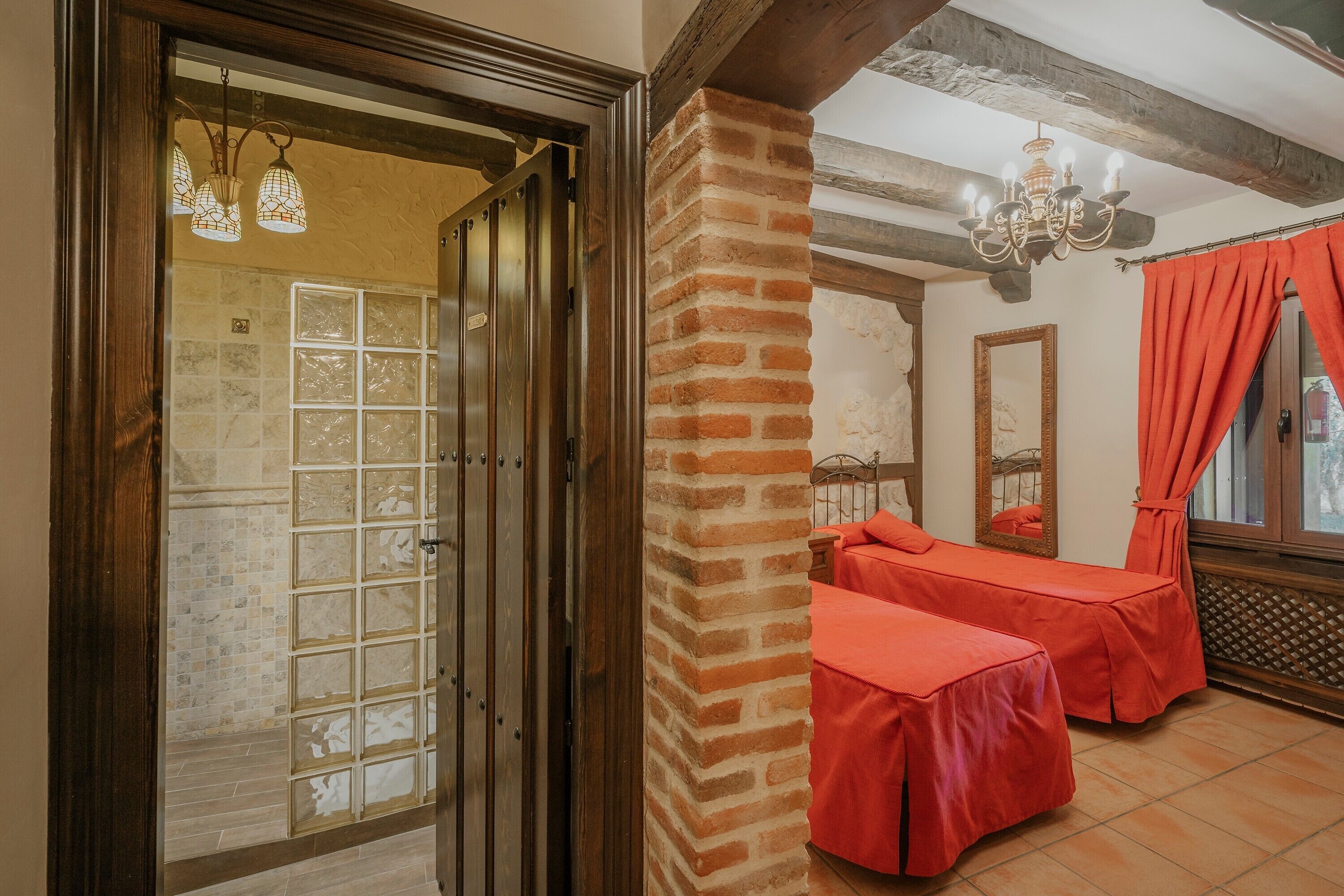 A room with a wooden door, a brick column, a chandelier, a mirror, and a window with orange curtains.