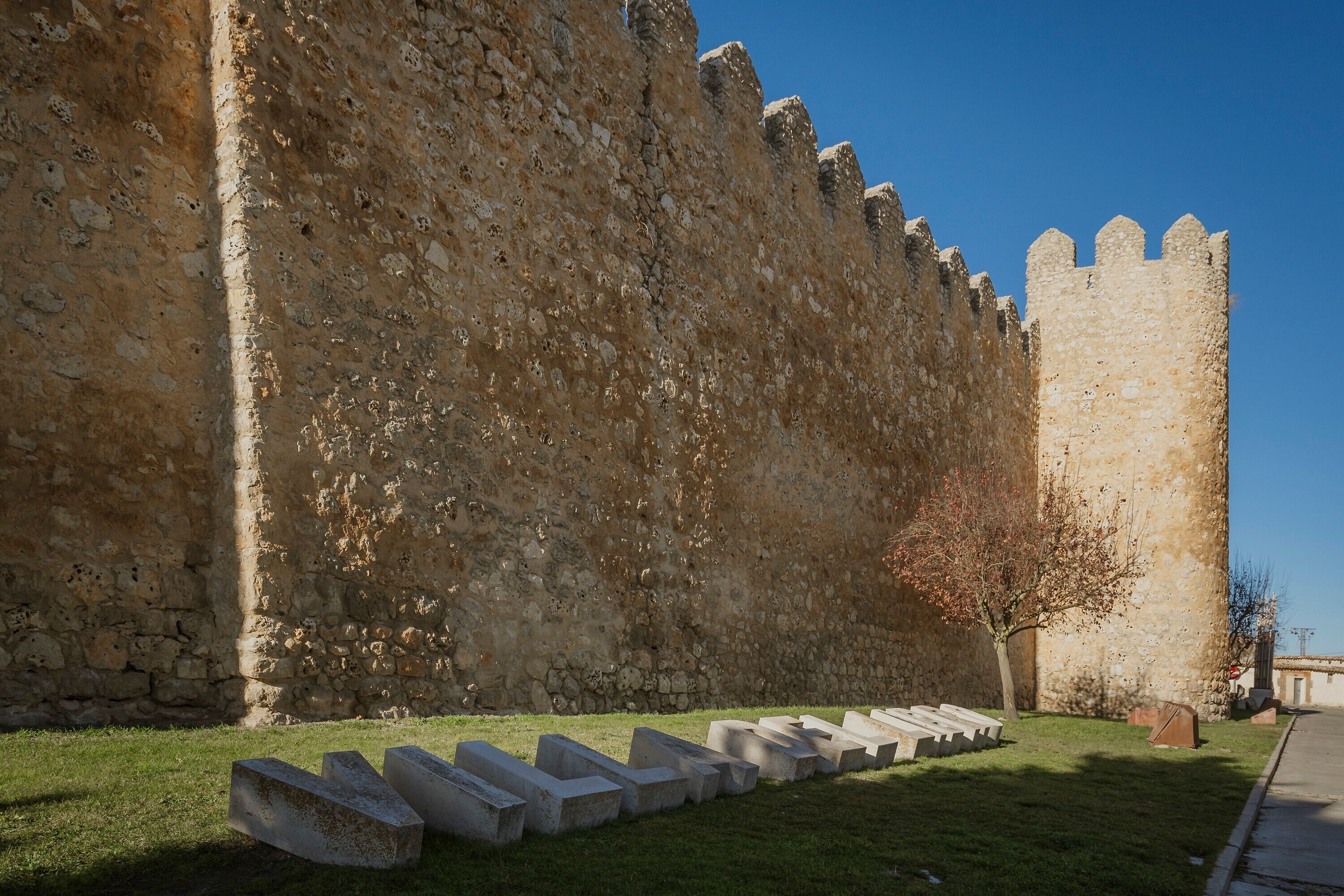 A stone wall with a tower, a grassy area with concrete benches, and a clear sky.