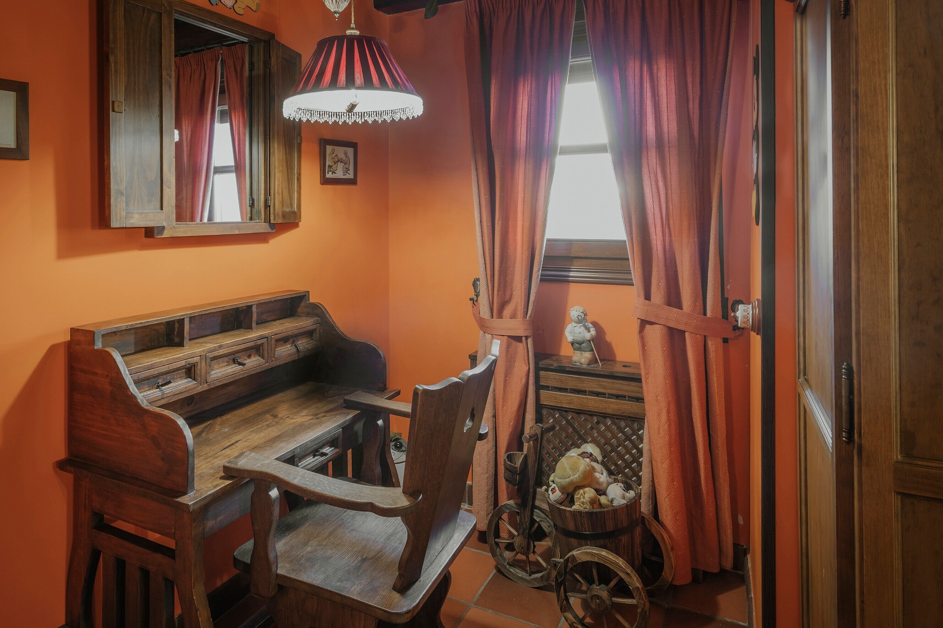Ein Zimmer mit einer hölzernen Bank, einem Stuhl, einem kleinen Tisch, einem Fenster mit Vorhängen und einer Wandlampe.