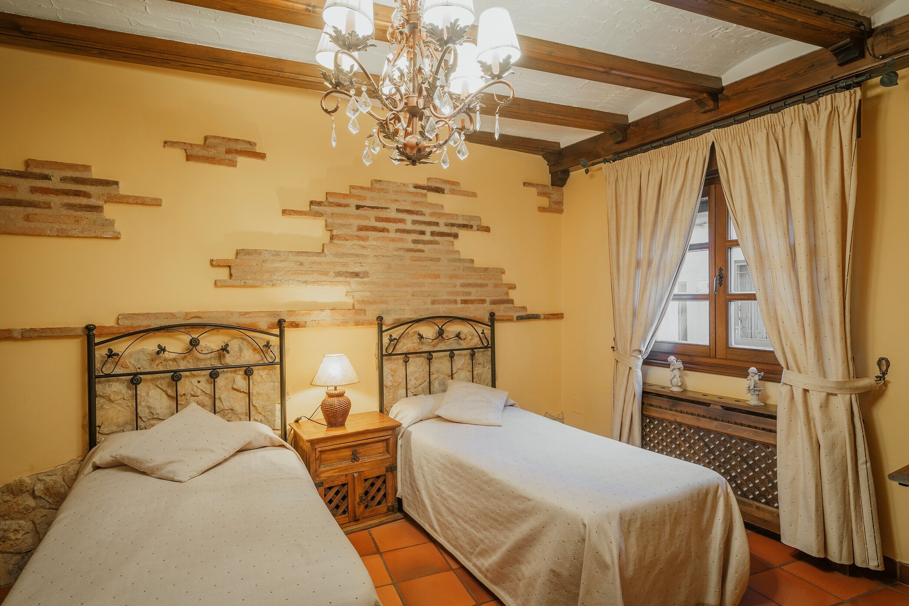 A room with two beds, a chandelier, a window with curtains, and a brick wall design.
