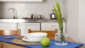 Een keuken met witte kastjes, een spoelbak en een eettafel met een blauwe placemat, een groene appel en een glazen vaas met een varen.