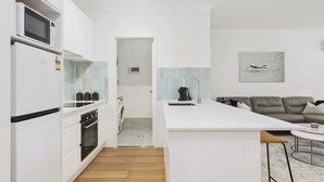 Uma cozinha moderna com armários brancos, forno embutido, micro-ondas e máquina de café. Há um sofá cinza e uma pequena mesa redonda na sala de estar.