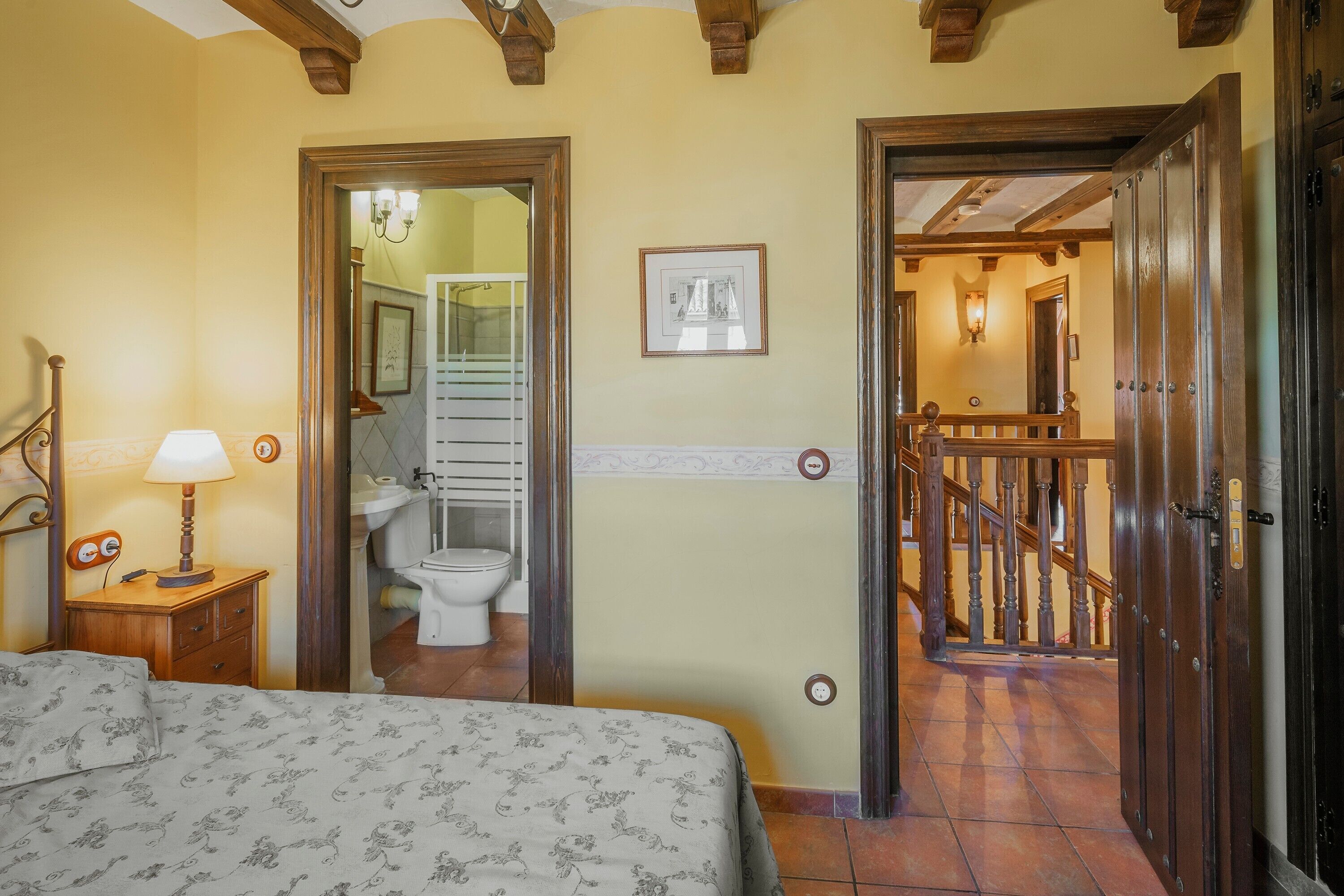 Un dormitorio con cama, una mesita de noche con lámpara, un baño con inodoro y lavamanos, y una escalera que lleva a otra habitación.
