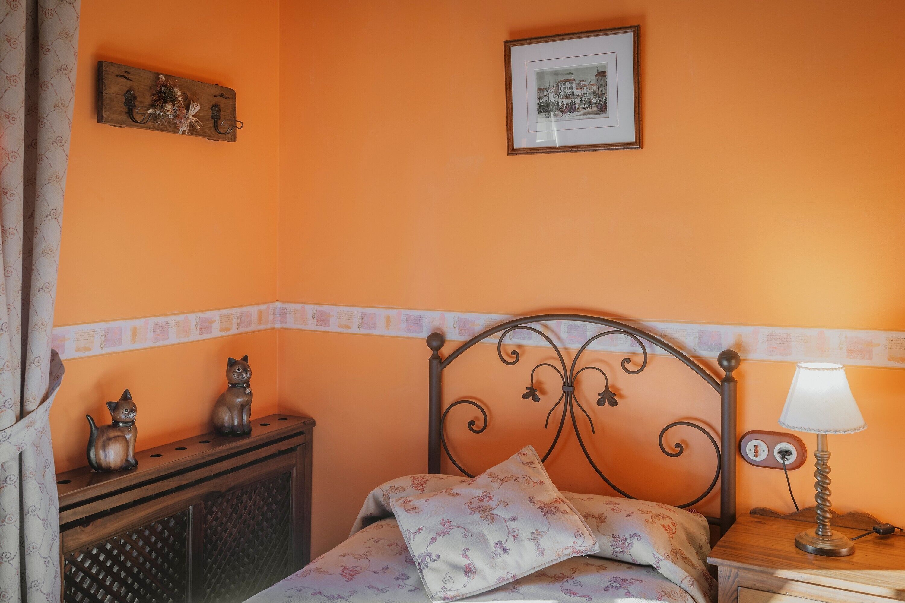 A bedroom with an orange wall, a bed with a floral bedspread, a nightstand with a lamp, and two cat figurines on a dresser.
