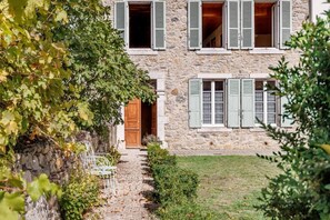 A stone building with a wooden door, multiple windows with shutters, and a paved path leading to the entrance.