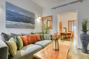 Een moderne woonkamer met een bank, een houten salontafel, een schilderij en een eethoek met stoelen en een tafel.