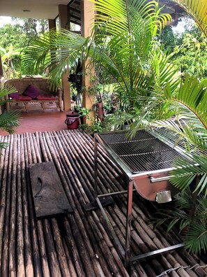 A covered outdoor area with a bamboo floor, a grill, a wicker sofa with red cushions, and various potted plants. - Mountain View House Kampot (Kampot)